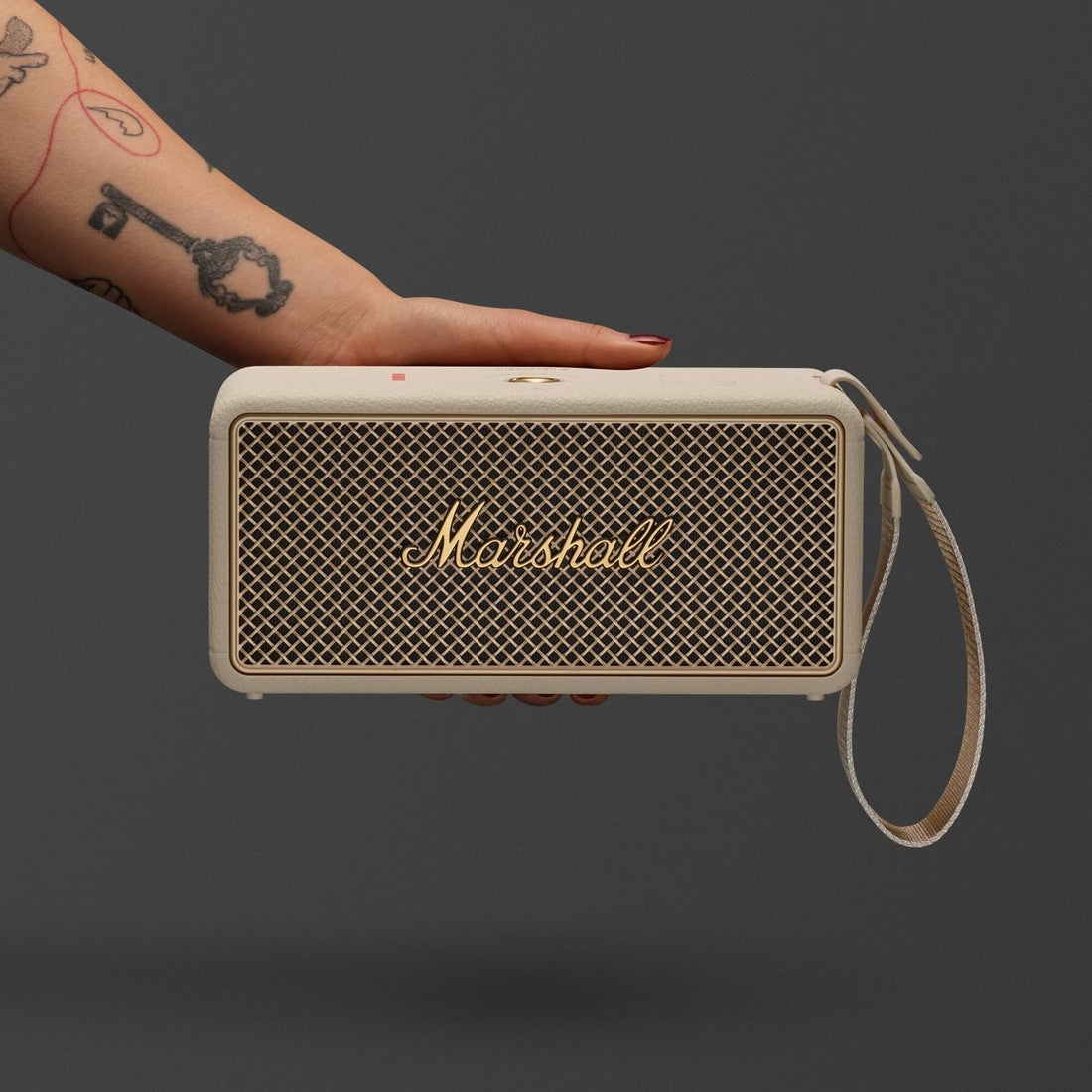 A tattooed hand holds the Marshall Middleton II Cream portable Bluetooth speaker against a plain dark gray background.