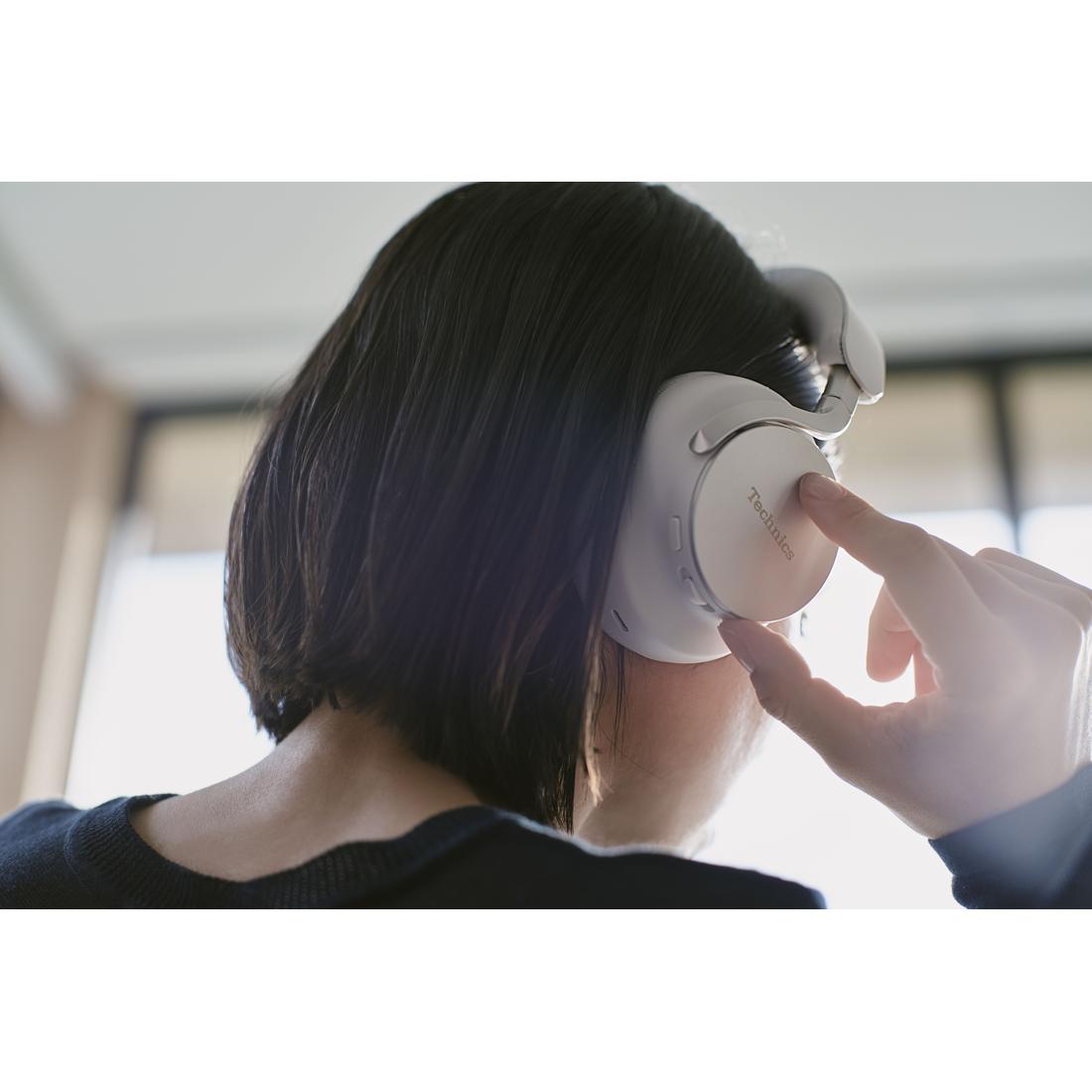 A person with short dark hair wearing white over-ear headphones and touching the earcup with a finger, shown from behind in a softly lit indoor setting.