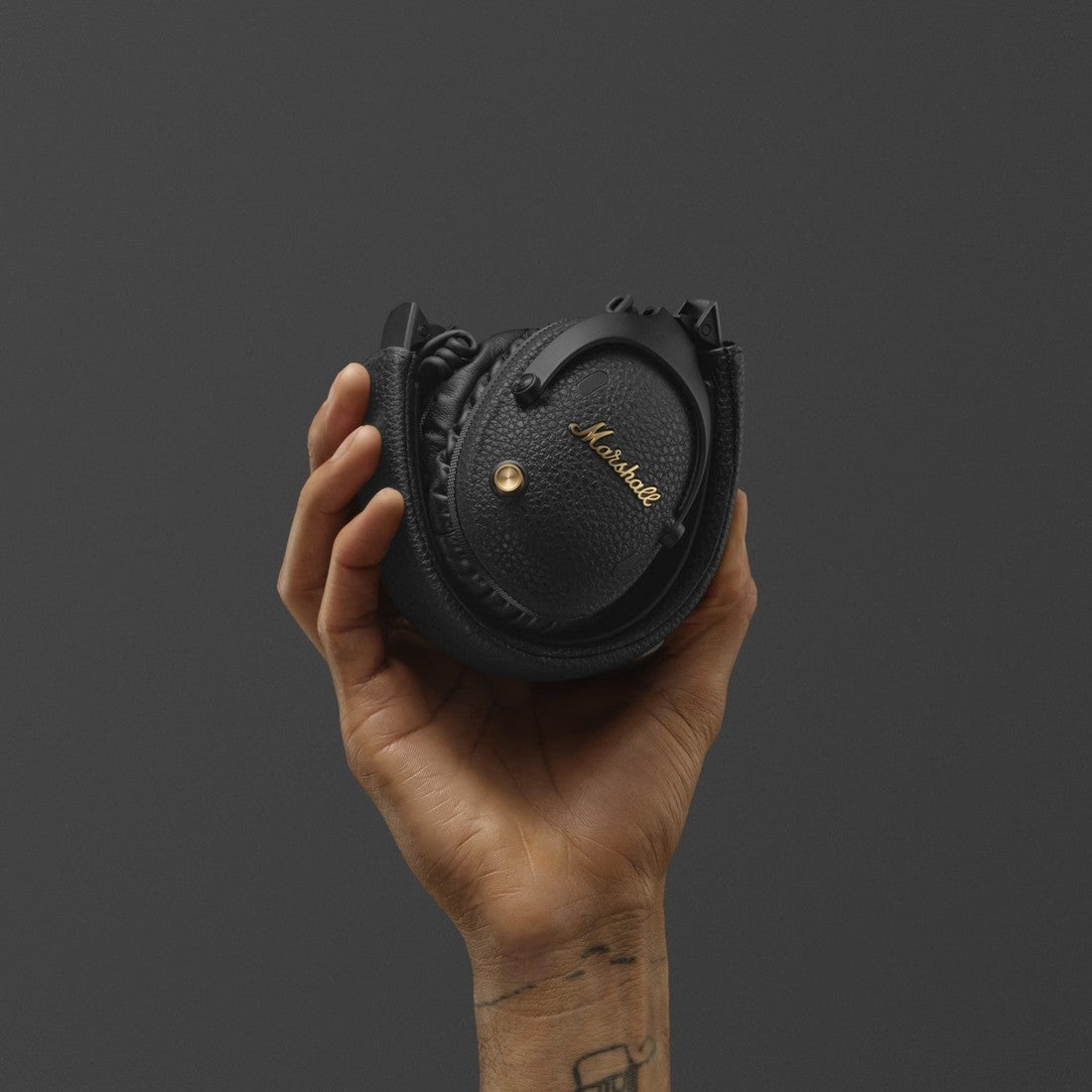 A folded pair of Marshall Monitor III A.N.C. Black bluetooth over-ear headphones in a hand against a dark gray background.