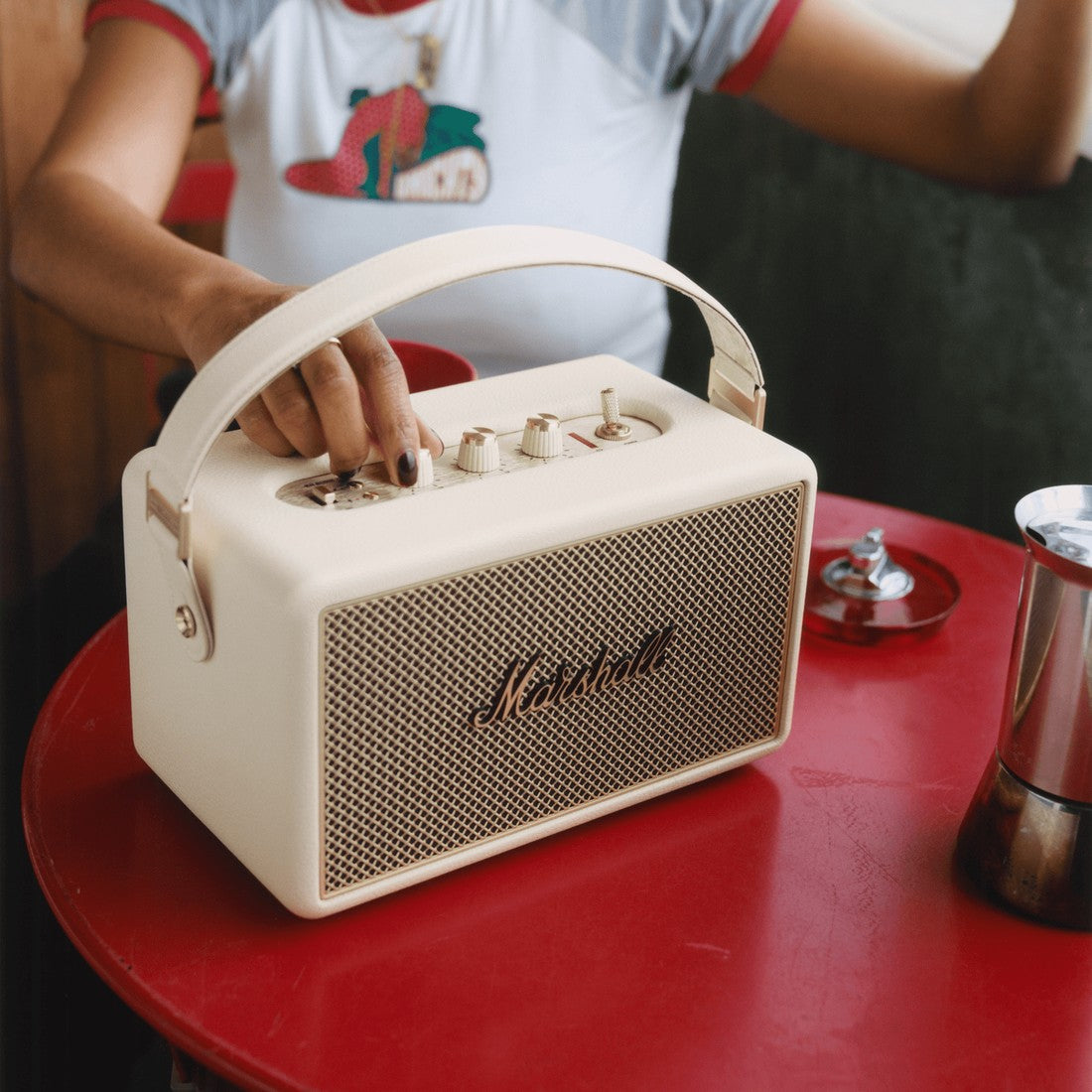 A person adjusts the knobs on a Marshall Kilburn III Cream portable speaker, set on a red table.