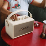 A person adjusts the knobs on a Marshall Kilburn III Cream portable speaker, set on a red table.