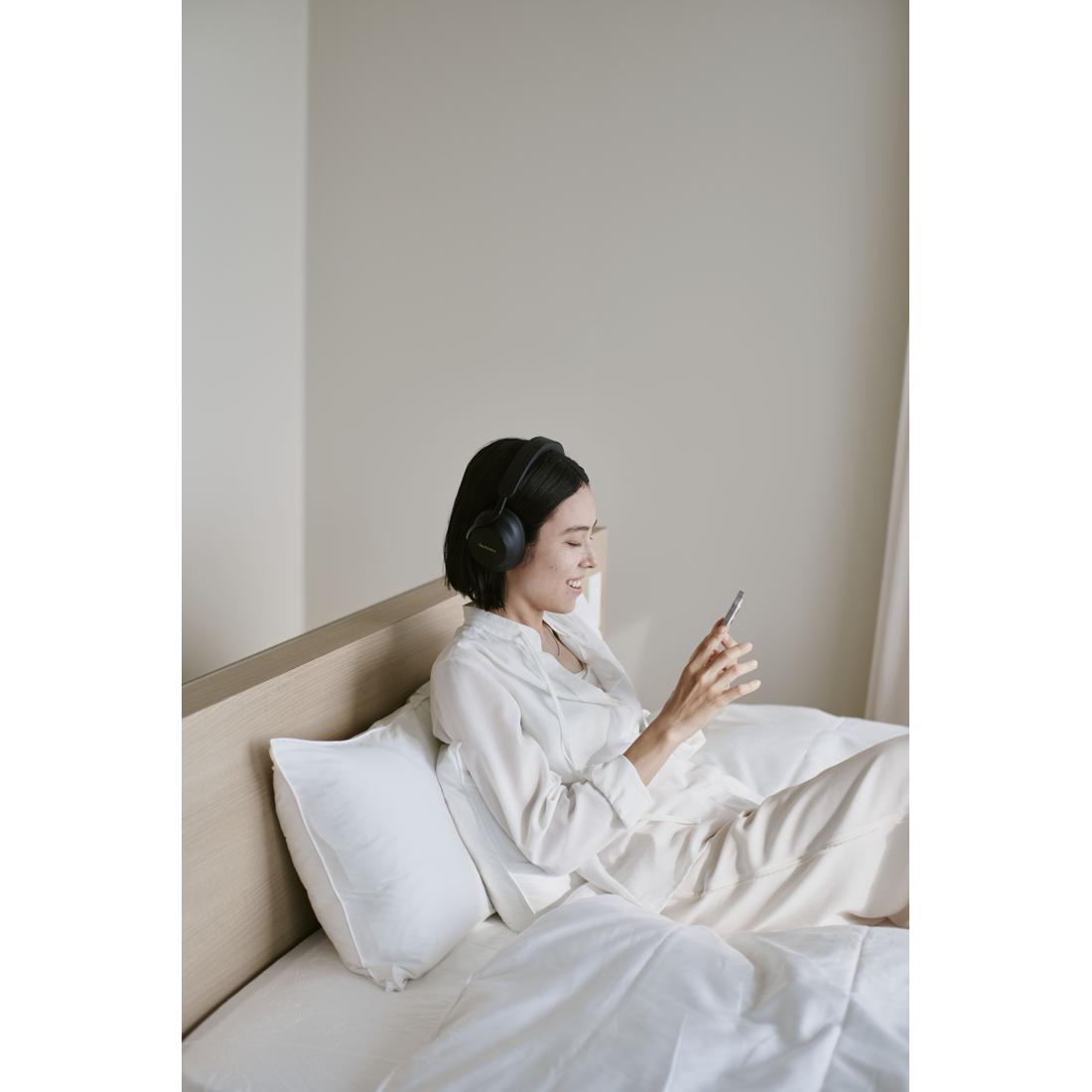 A woman wearing white pajamas sits on a bed with white pillows and bedding, wearing over-ear headphones and looking at her phone in a softly lit bedroom.