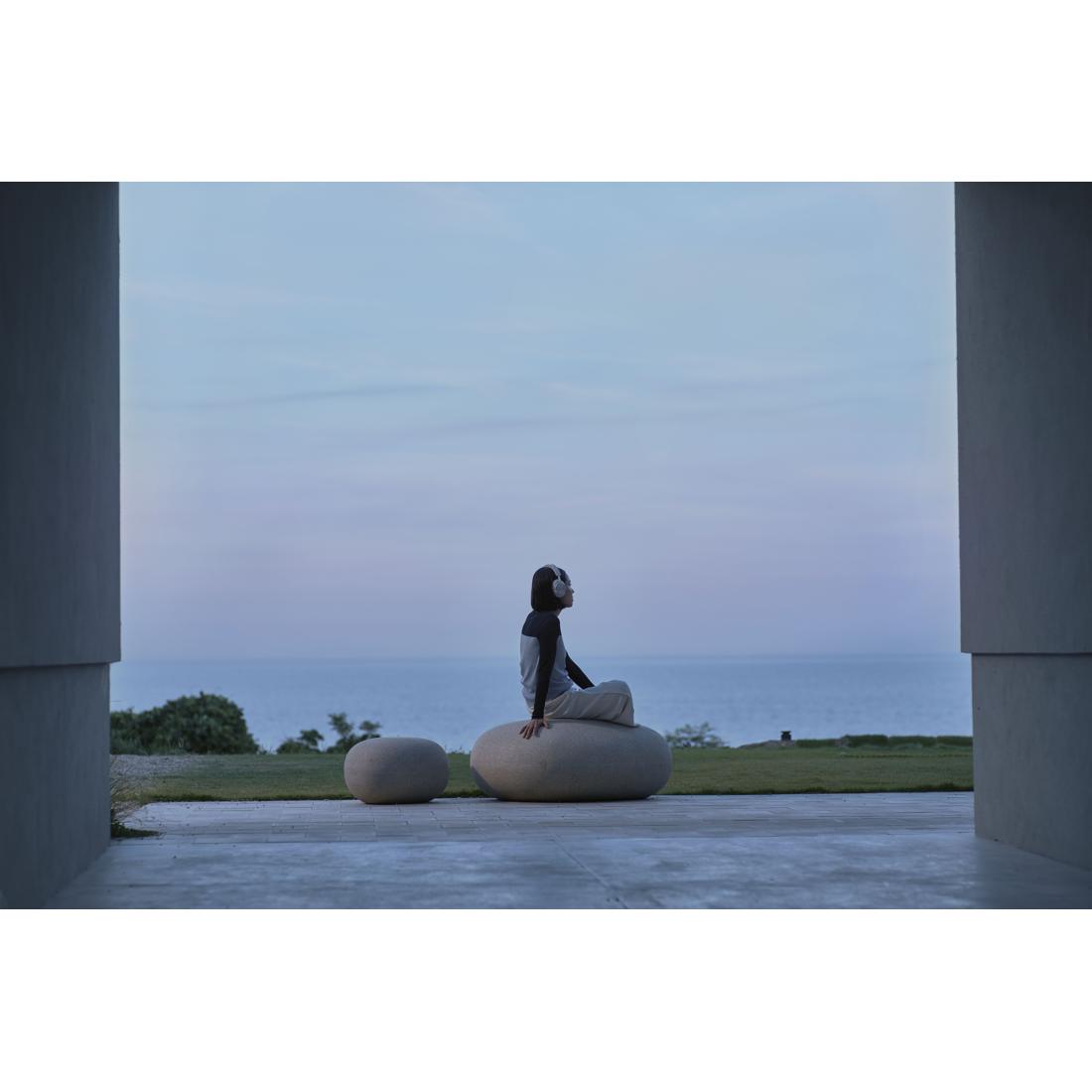 A person sits cross-legged on a large oval stone bench outdoors, framed by two tall columns, with a calm sea and pale sky in the background.