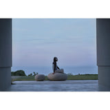A person sits cross-legged on a large oval stone bench outdoors, framed by two tall columns, with a calm sea and pale sky in the background.