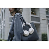 A man in a dark gray oversized jacket carries a large dark gray tote bag over his shoulder, with white over-ear headphones draped over the bag in an outdoor urban setting.