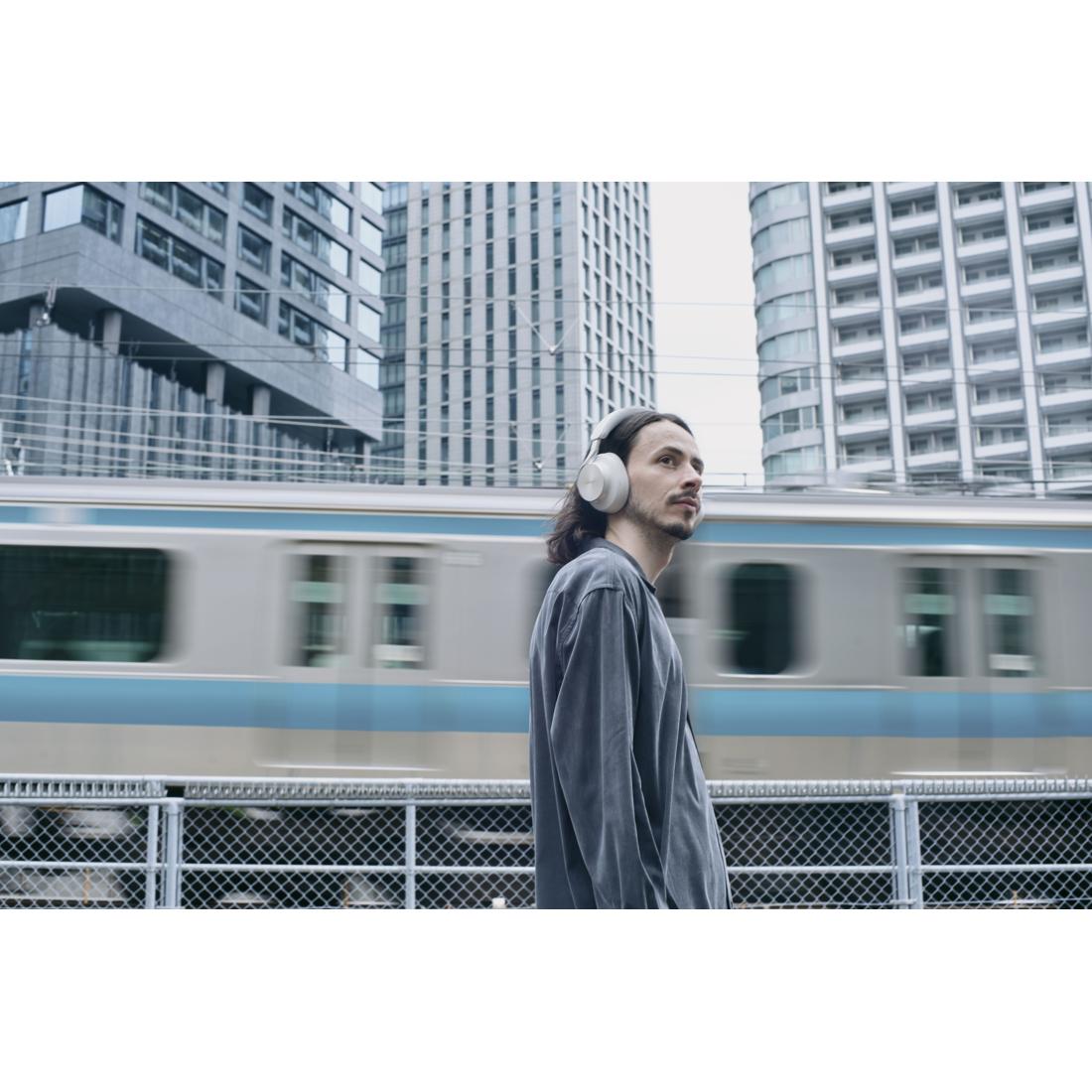 A man wearing white over-ear headphones stands in an urban setting with a passing train and tall buildings in the background.