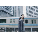 A man wearing white over-ear headphones stands in an urban setting with a passing train and tall buildings in the background.