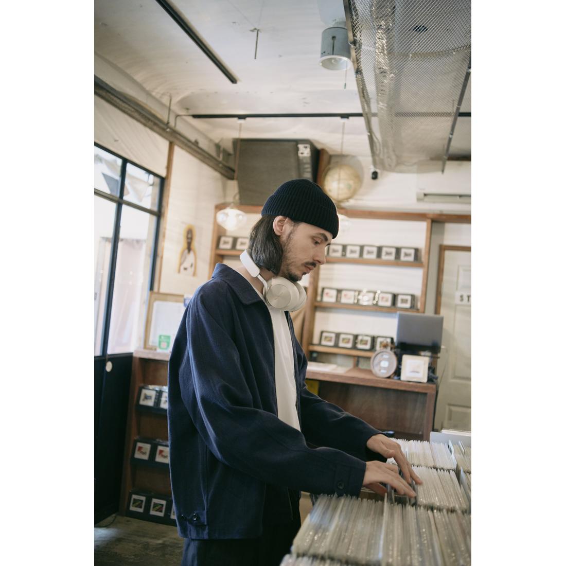 A man wearing a black beanie, navy blue jacket, and white headphones around his neck is seen in a vintage record store, looking down at a stack of vinyl records.