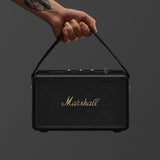 A tattooed hand holds the Marshall Kilburn III Black and Brass portable speaker by its handle against a plain background.