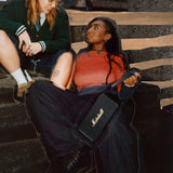 Two people sit on stairs, one holding a Marshall Middleton II Black and Brass portable Bluetooth speaker.