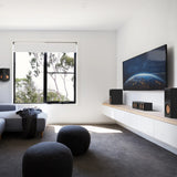 A modern home theater setup in a bright, minimalist room. The scene features a large wall-mounted television displaying Earth, flanked by black speakers on a floating light-wood shelf with white cabinets.