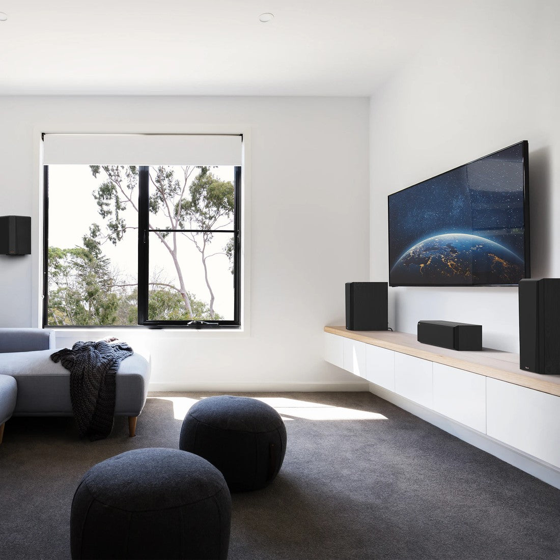 A modern living room setup featuring a large flat-screen TV mounted on a white wall above a floating shelf with speakers, a gray sofa, and two dark round ottomans on a dark gray rug.
