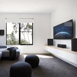 A modern living room setup featuring a large flat-screen TV mounted on a white wall above a floating shelf with speakers, a gray sofa, and two dark round ottomans on a dark gray rug.