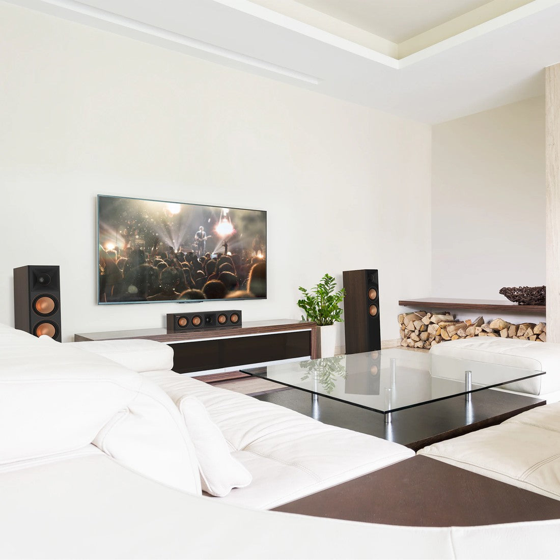 A modern living room setup featuring a large flat-screen TV mounted on a white wall above a dark wood console, flanked by two walnut-finish Klipsch speakers. In the foreground, a white sectional sofa and a glass-top coffee table are visible.