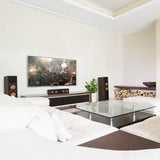 A modern living room setup featuring a large flat-screen TV mounted on a white wall above a dark wood console, flanked by two walnut-finish Klipsch speakers. In the foreground, a white sectional sofa and a glass-top coffee table are visible.