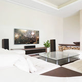 A modern living room interior with a large wall-mounted television displaying a concert scene, set above a low media console. The room features white upholstered seating, a glass-top coffee table with dark wood framing, walnut-finish Klipsch speakers, and a built-in wood storage niche with stacked firewood.