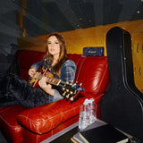 A woman sits on a red couch playing an electric guitar. Nearby are a guitar case, two water bottles, a notebook, and a Marshall Emberton III Hendrix Edition speaker.
