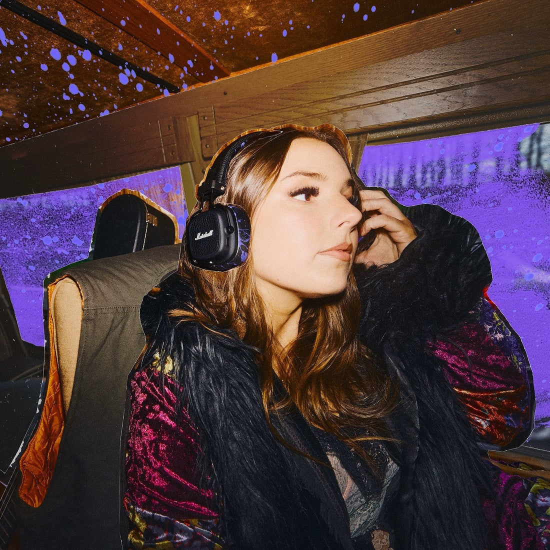 A woman wearing Major V Hendrix Edition headphones sits on a bus with purple lighting and raindrops on the window behind her.