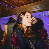 A woman wearing Major V Hendrix Edition headphones sits on a bus with purple lighting and raindrops on the window behind her.