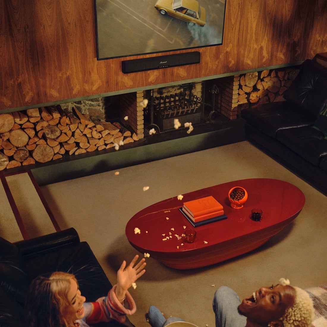 Two people sitting on a couch in a living room with popcorn and a TV playing a car video, with a Marshall Heston 60 mounted to the wall